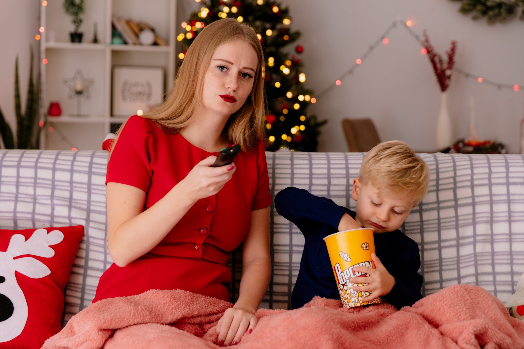 happy mother red dress with her little child sitting couch blanket with bucket popcorn watching tv together decorated room with christmas tree wall